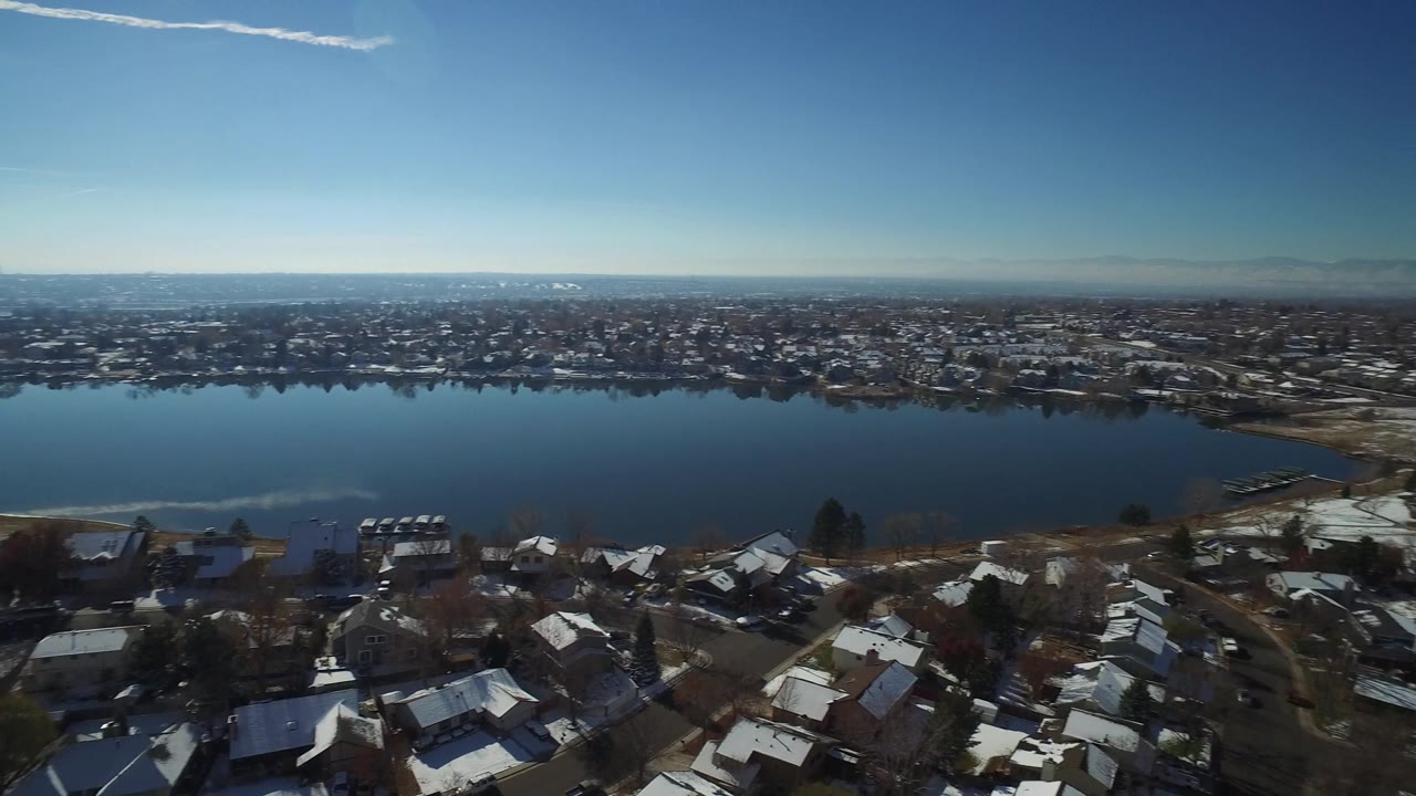 Wide aerial of a lake, residential shoreline, and town spread toward mountains.