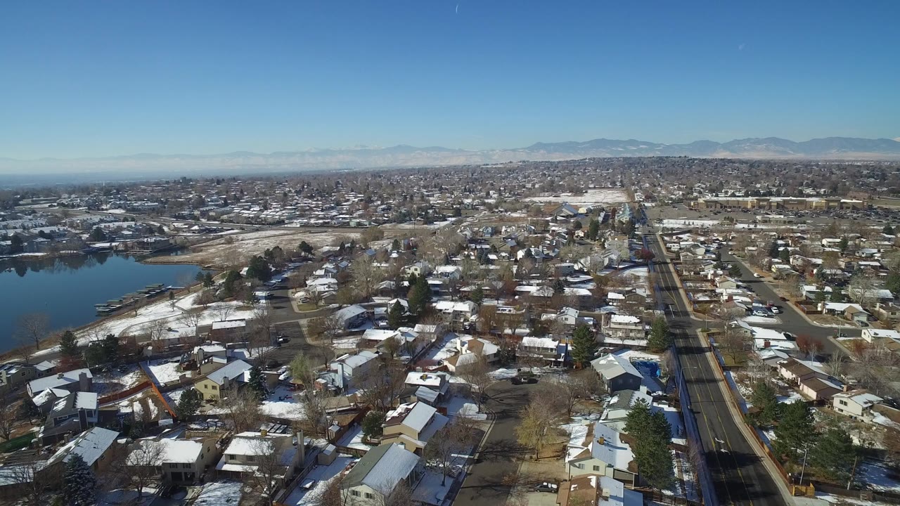 Aerial view over snowy homes, roads, lake edge, and distant mountains.