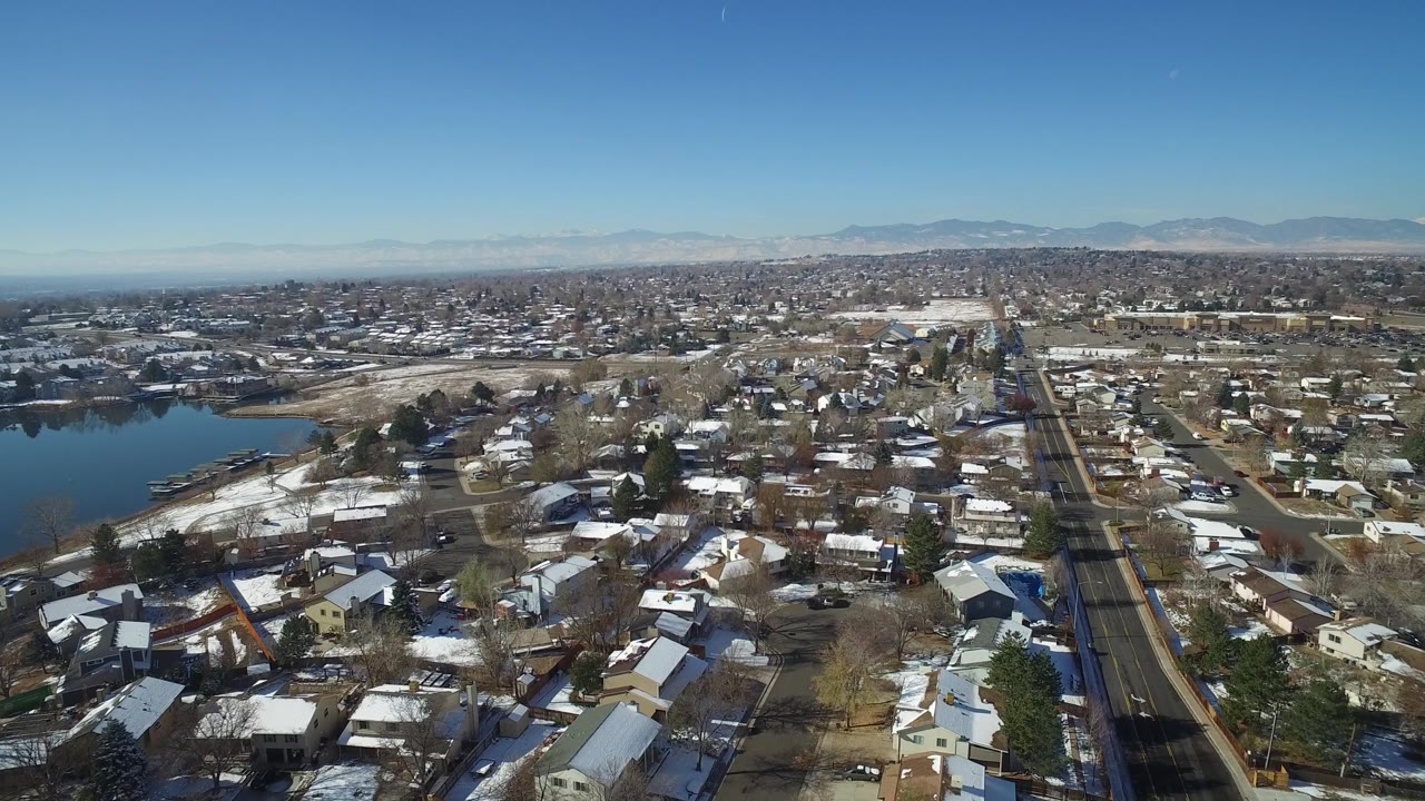 Aerial frame over Hidden Lake, Colorado with clustered homes, patchy snow, a road grid, the lake partly visible on the left, a hazy mountain backdrop, and no visible people.