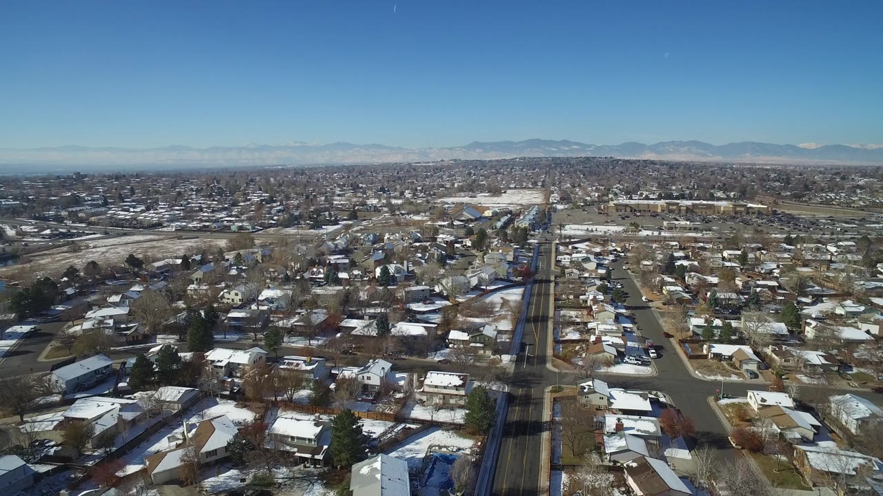 Second aerial frame over Hidden Lake, Colorado, with a long road corridor near center, compact homes on both sides, winter snow patches, and no visible people.