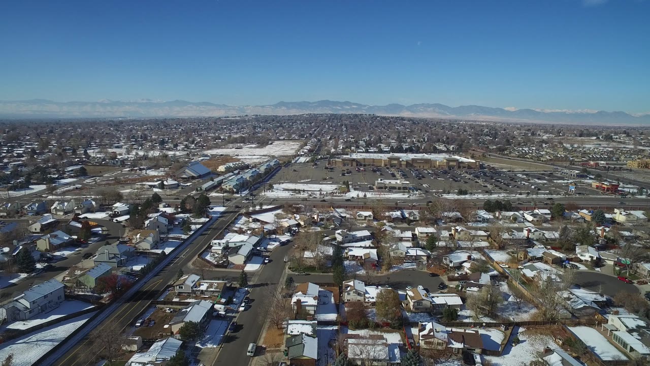 Hidden Lake, Colorado appears from above with a diagonal arterial road at left, houses and yards below, open paved areas in the midground, mountains, and no visible people.