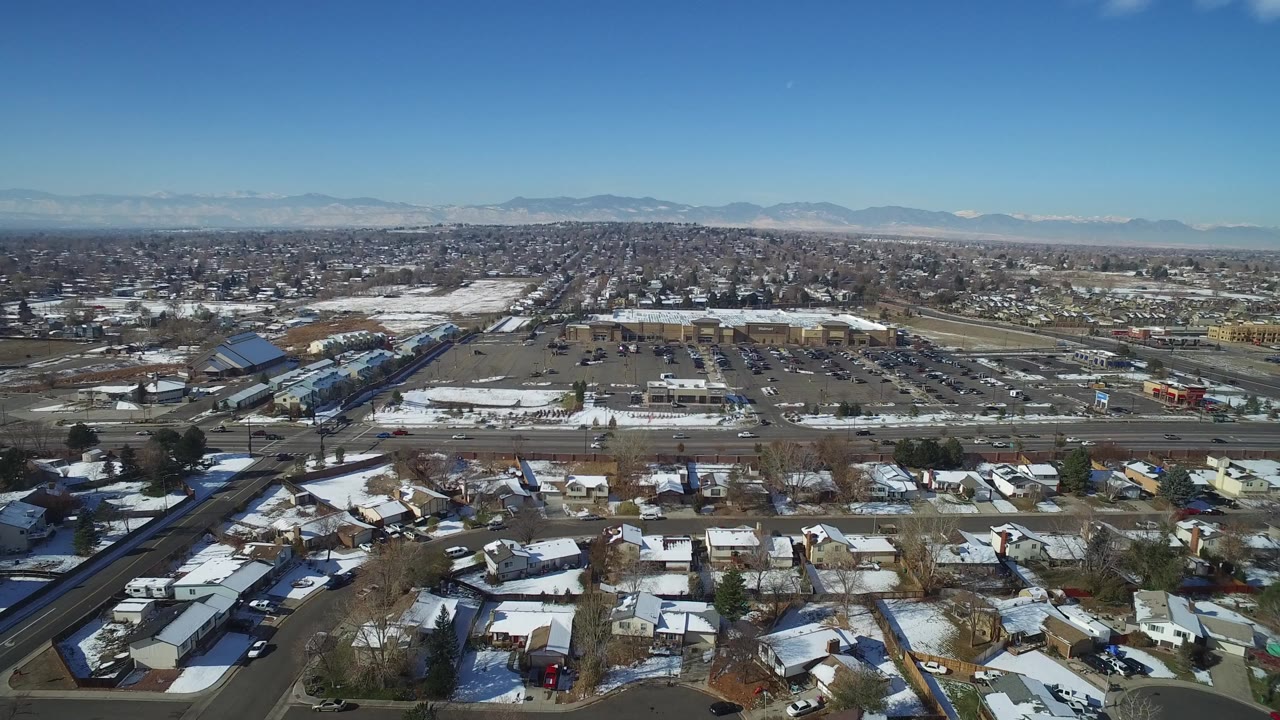 Wide aerial view of snowy houses, roads, parking lots, and distant mountains.