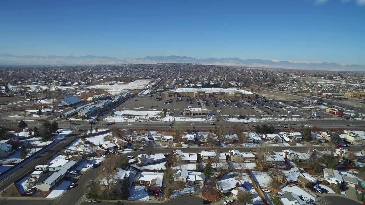 Elevated drone frame of Hidden Lake, Colorado showing neighborhood rooftops at the bottom, a multi-lane road with cars, large parking areas, low buildings, distant houses, mountains, and no visible people.
