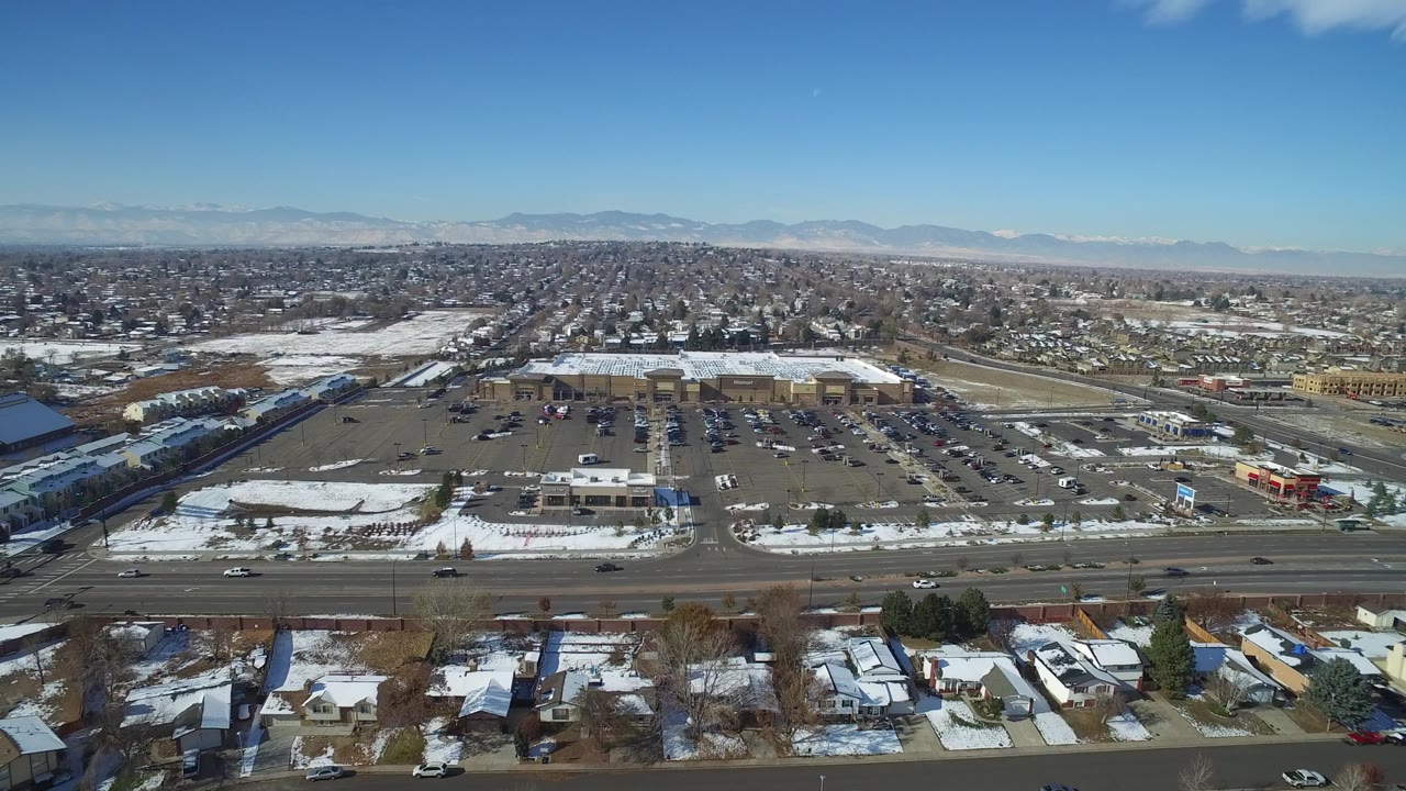 Wide aerial frame in Hidden Lake, Colorado with houses in the foreground, a multi-lane road, retail parking areas, snow-covered ground, distant mountains, and no visible people.