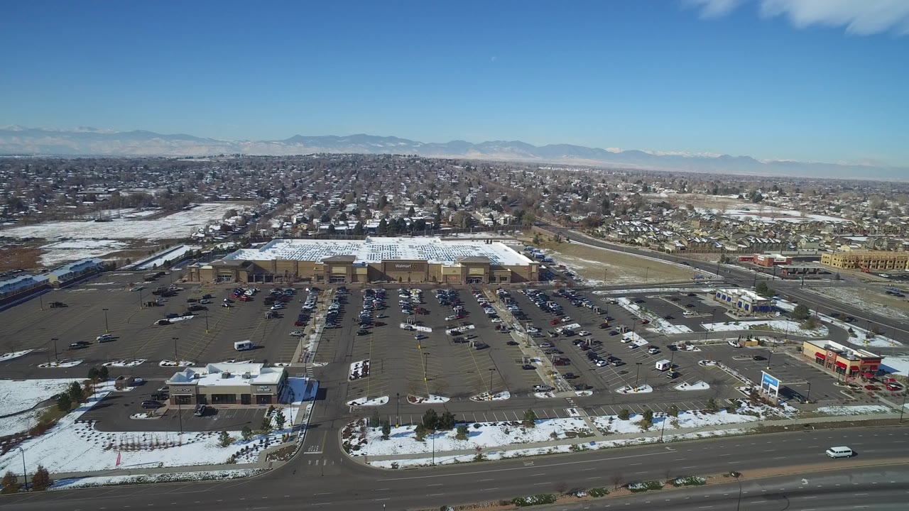 Aerial view of a mall parking lot, nearby roads, houses, snow patches, and distant mountains.