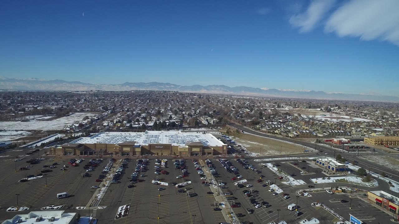 Aerial view of a shopping center parking lot, snowy roofs, suburbs, and distant mountains.