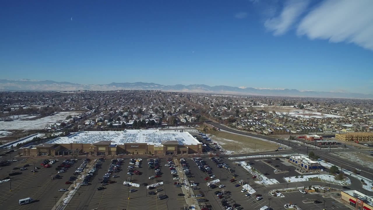 Aerial view of snowy retail buildings, parking lots, roads, houses, and distant mountains.