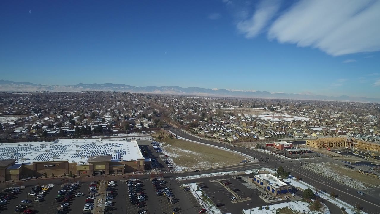 Aerial view of suburban roads, retail buildings, homes, snow patches, and distant mountains.