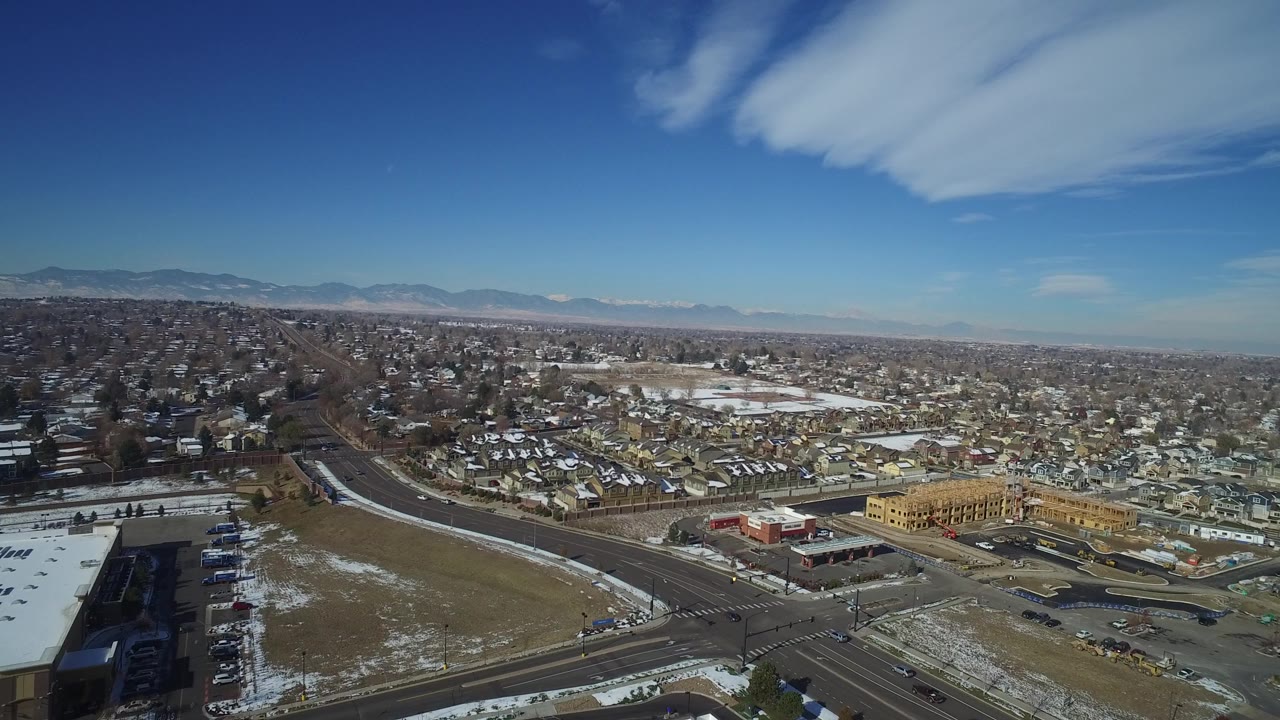 Aerial view of suburban streets, houses, commercial buildings, patchy snow, and distant mountains.
