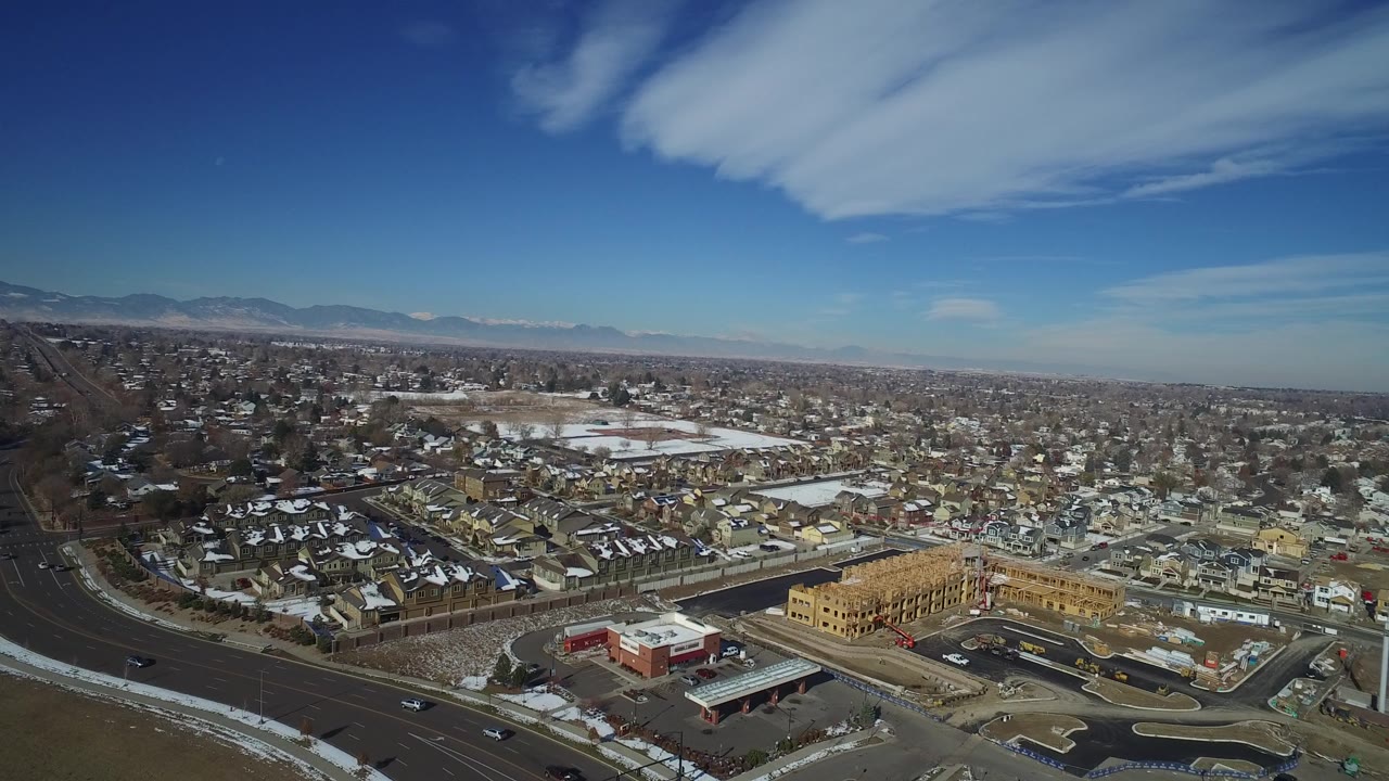 High aerial frame over Hidden Lake, Colorado, with a tan construction site near the foreground, snow-dusted homes, roads, distant mountains, blue sky, and no visible people.
