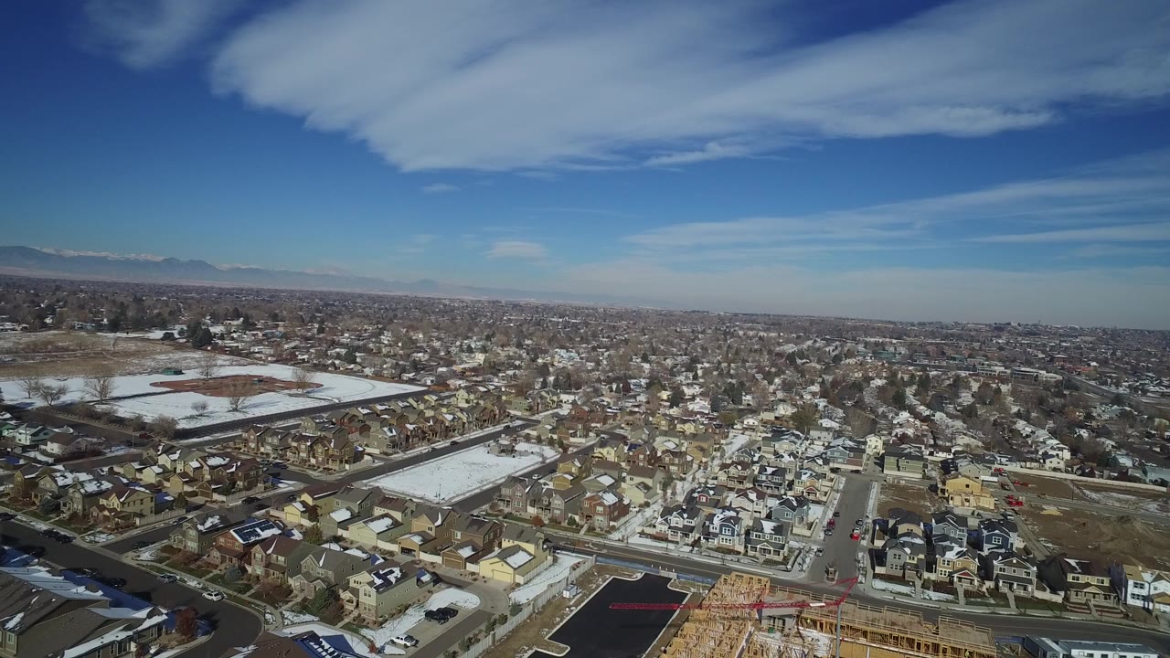 Aerial view of suburban homes, roads, parking lots, snow patches, and distant mountains.