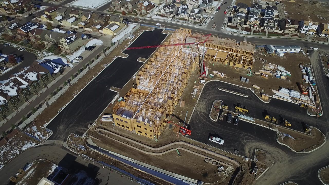 High oblique aerial frame of a Hidden Lake, Colorado residential construction site with wood framing, paved roads, nearby houses, snow patches, and no visible people.