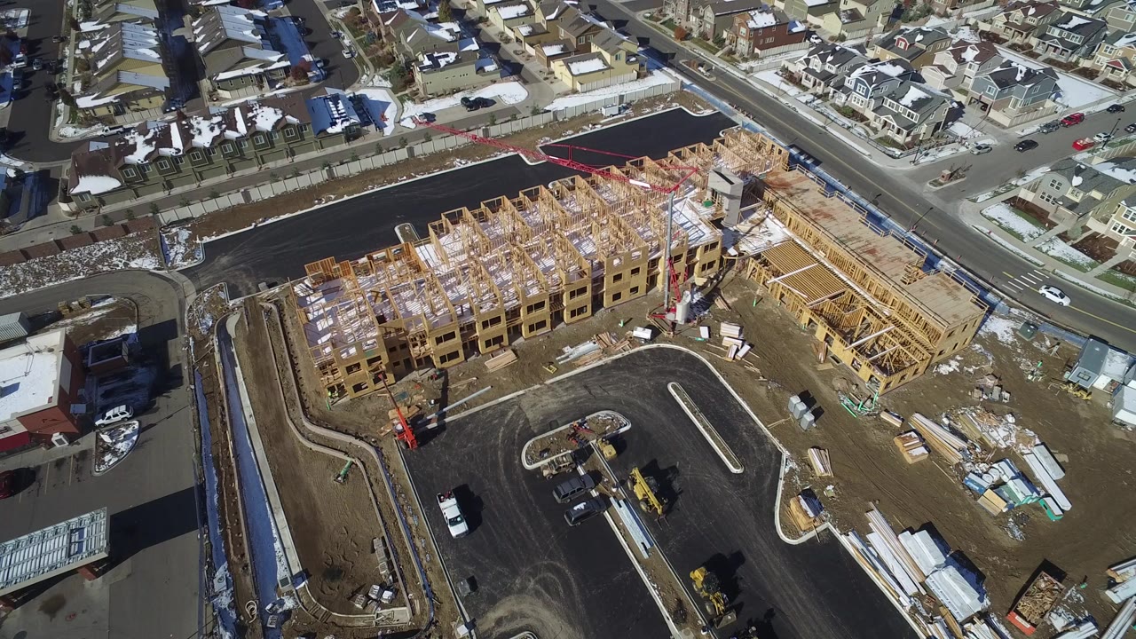 Aerial frame from Hidden Lake, Colorado showing wood-framed buildings under construction, a curved paved road, parked equipment, nearby houses, snow patches, and no visible people.