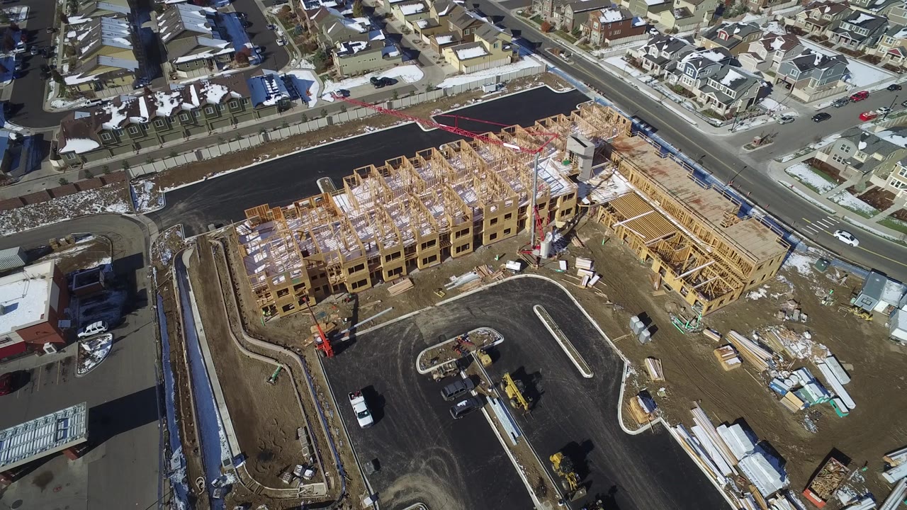 Aerial view in Hidden Lake, Colorado with the framed structure centered, asphalt lanes and curbs below, houses and a roadside commercial building around the site, and no visible people.