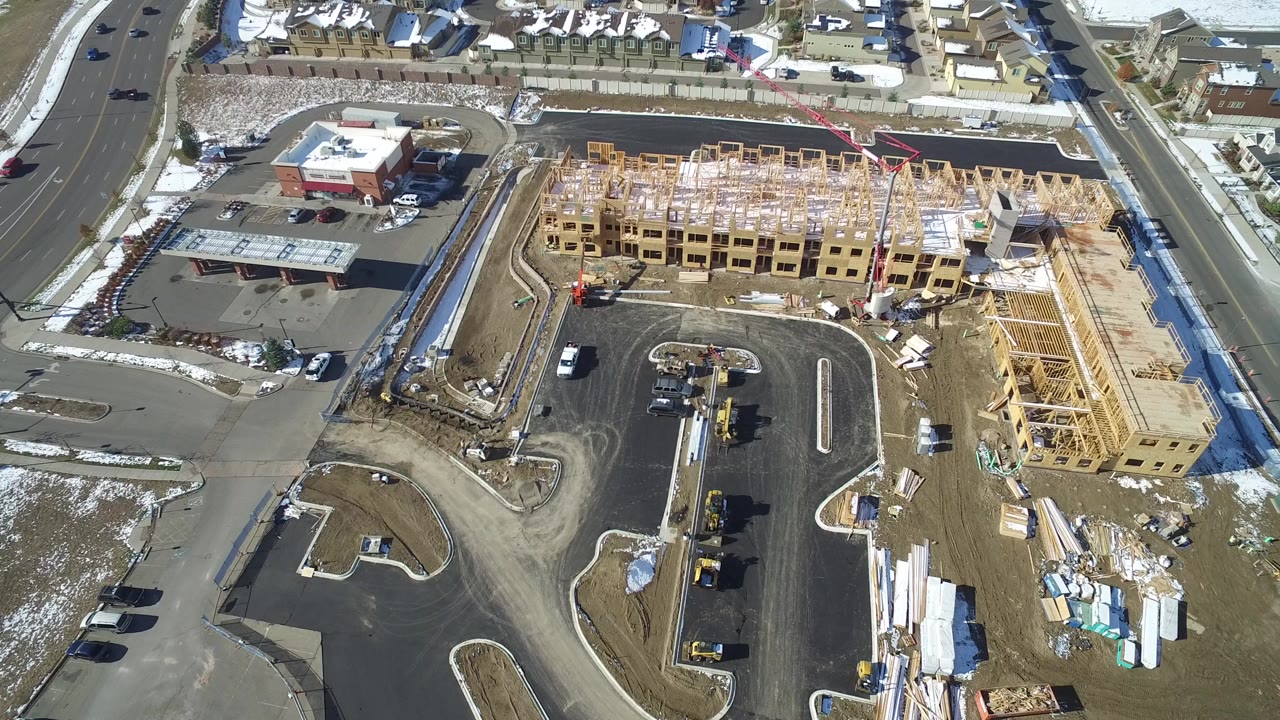 Aerial frame over Hidden Lake, Colorado showing a wood-framed building under construction, paved roads, a parking lot, construction materials, snow patches, and no visible people.