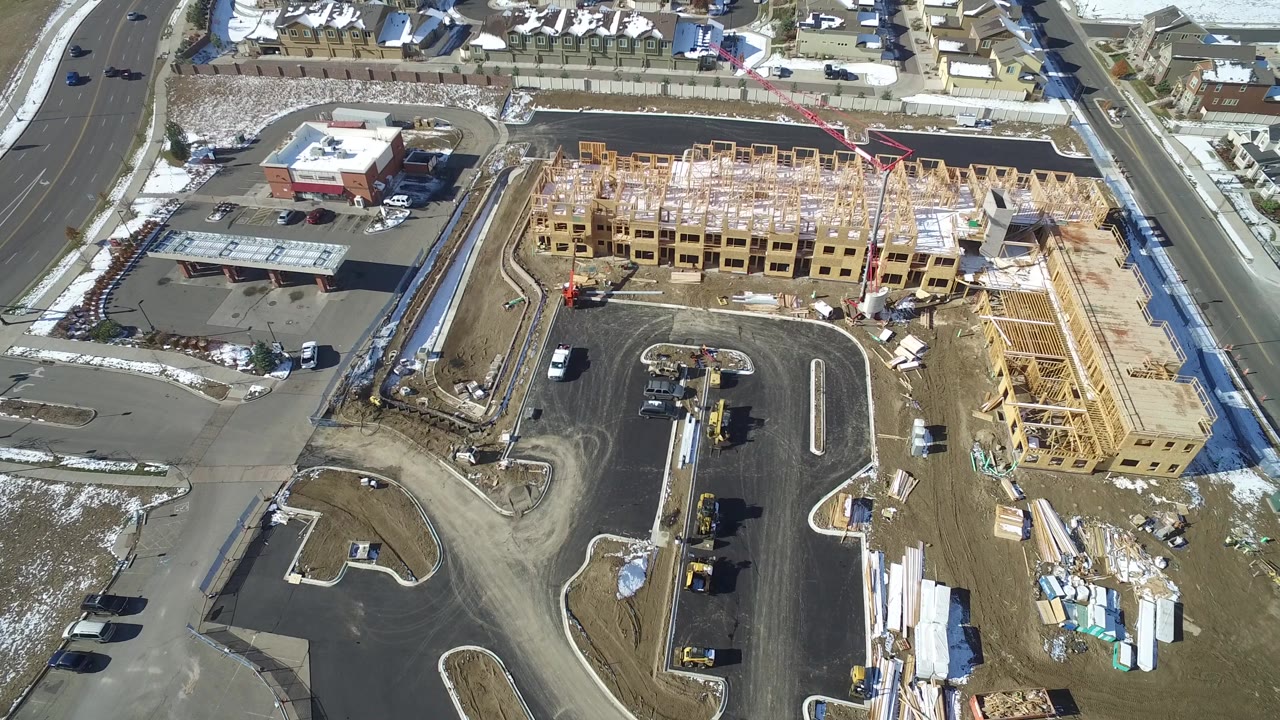 Overhead construction site with exposed timber framing, paved streets, and residential rooftops nearby.
