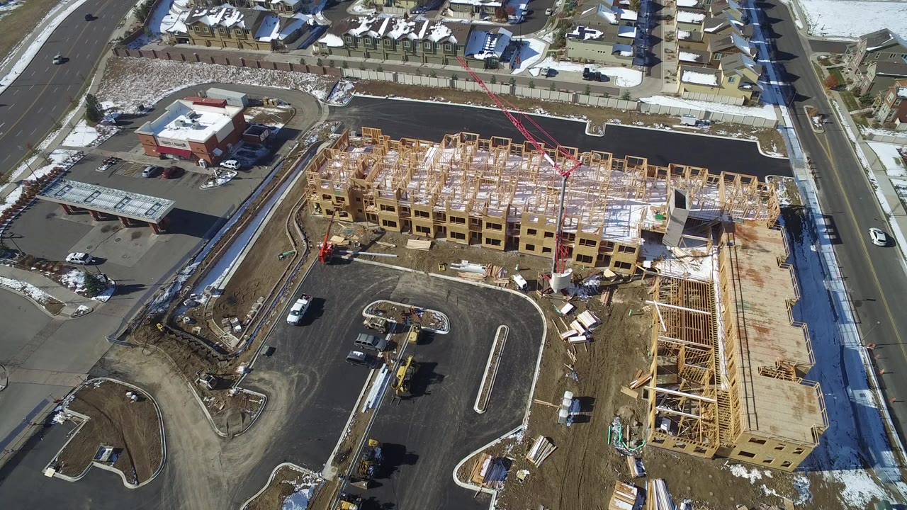 High aerial view over Hidden Lake, Colorado, showing a large wood-framed building site beside paved roads, a parking area, snow patches, nearby homes, and no visible people.