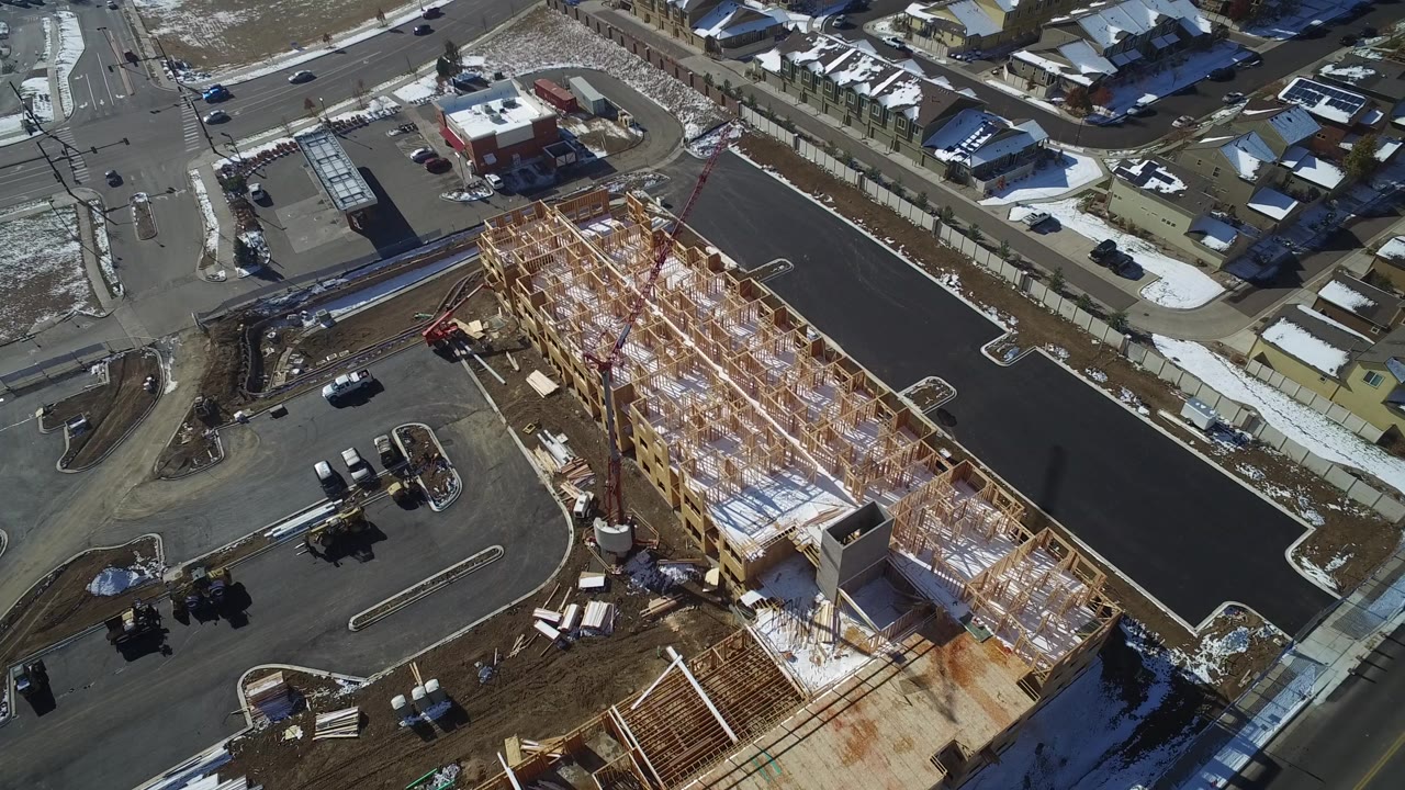 Aerial frame over Hidden Lake, Colorado showing a large wood-framed building under construction beside paved roads, parked vehicles, snow patches, and nearby houses, with no visible people.