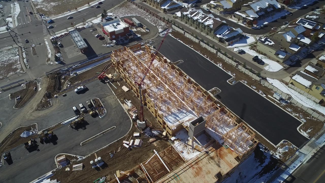 Overhead view in Hidden Lake, Colorado with exposed timber framing across a long construction footprint, dark asphalt roads, parked cars, sidewalks, snow, and residential houses, with no visible people.