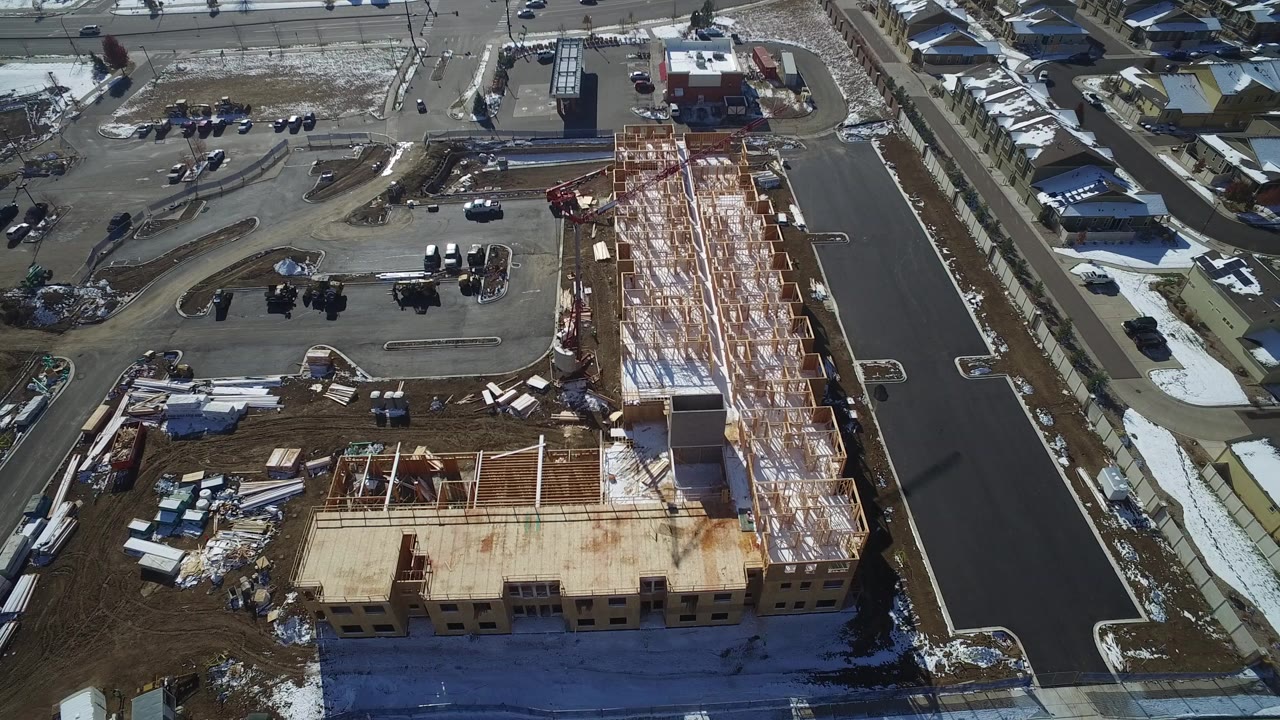 Overhead frame in Hidden Lake, Colorado showing an L-shaped wood-framed building under construction beside dark paved streets, snow patches, parked vehicles, nearby houses, and no visible people.