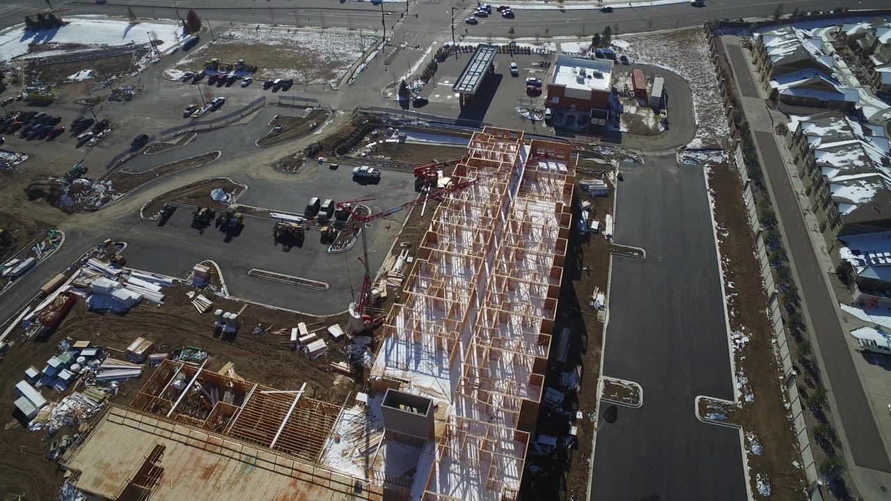 Aerial view in Hidden Lake, Colorado shows roof trusses across a long building, a red boom lift near the work area, parked cars, material stacks, and a road along the top of frame.