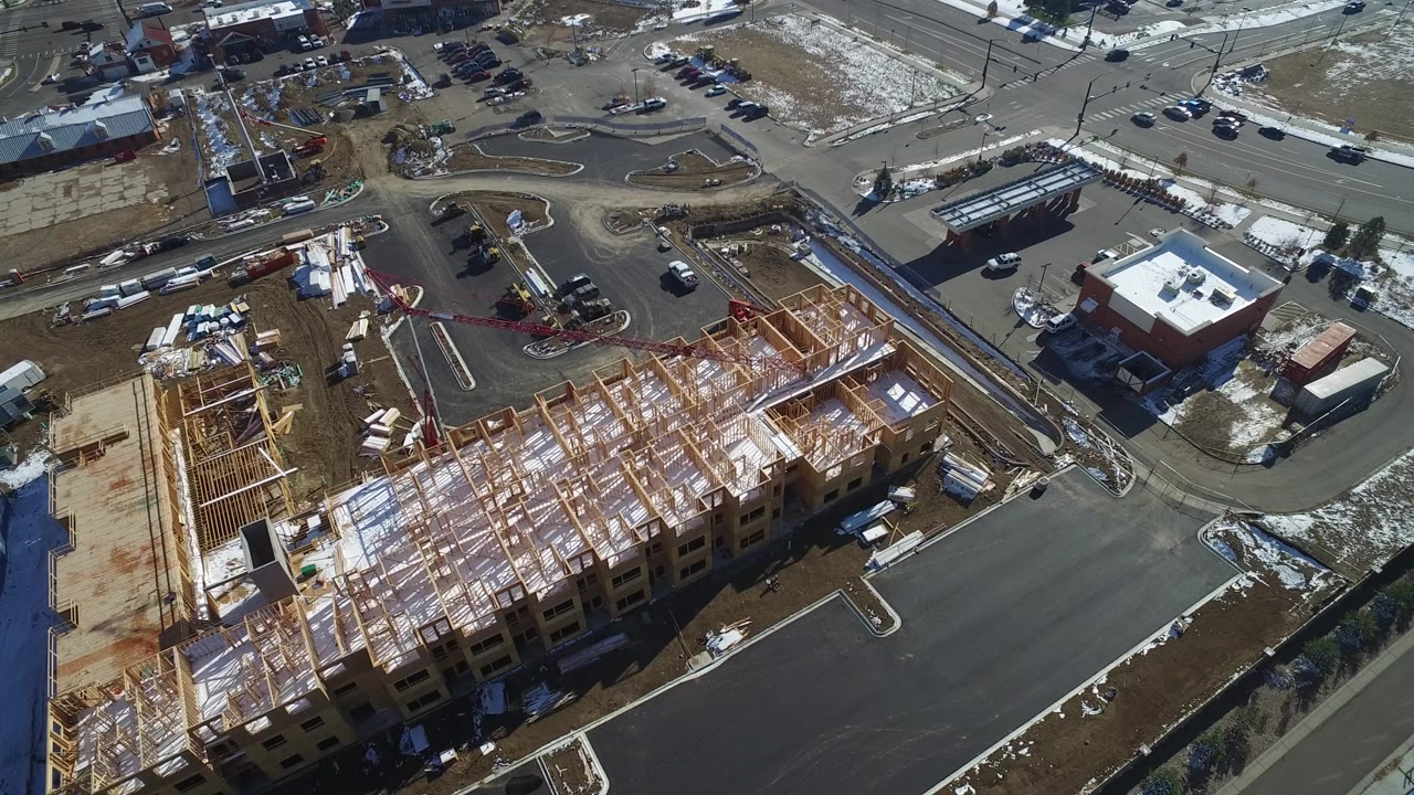 High aerial frame over Hidden Lake, Colorado, showing a wood-framed building under construction, a dark asphalt parking lot, surrounding streets, patchy snow, and no visible people.