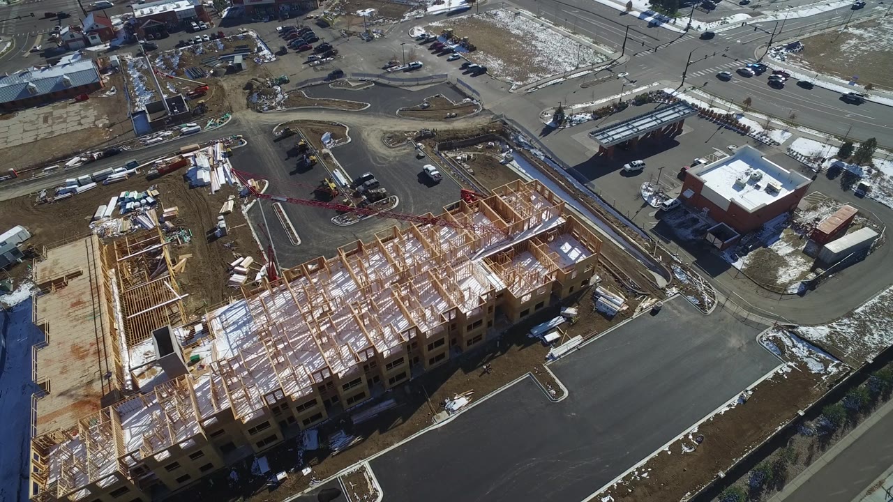 Aerial view in Hidden Lake, Colorado, with the unfinished timber structure centered below, parking lot pavement in front, road intersections above, scattered vehicles, and no visible people.