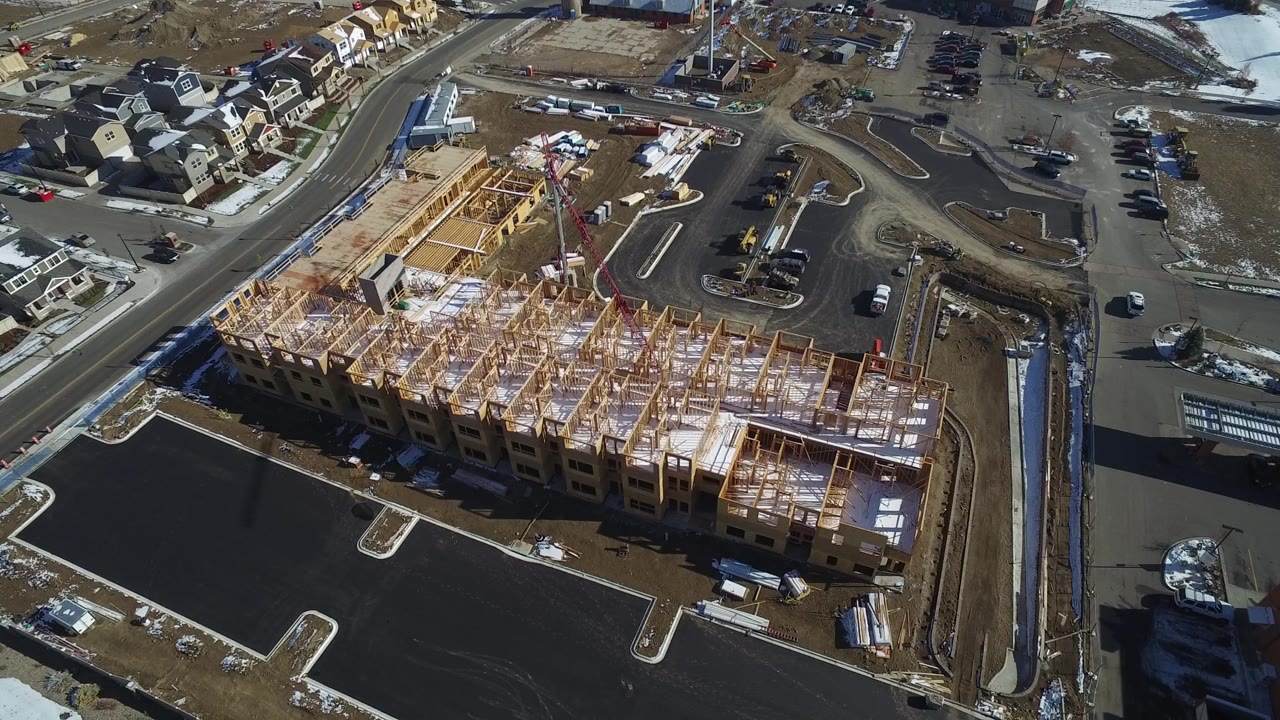 Aerial frame over Hidden Lake, Colorado showing a large wood-framed building under construction beside dark paved parking areas, dirt roads, nearby houses, and no visible people.