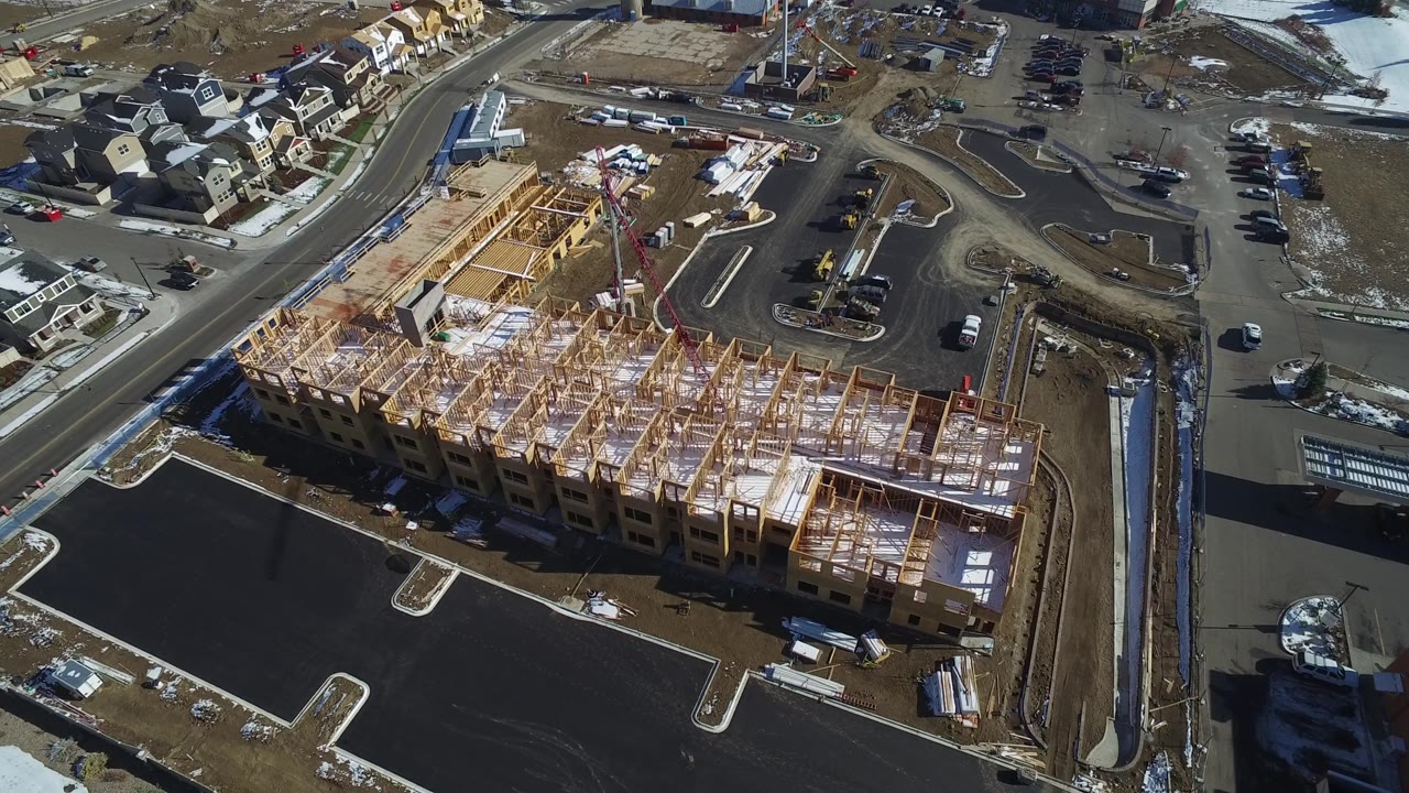 Second aerial frame in Hidden Lake, Colorado showing the unfinished building from above, with fresh asphalt, curved access roads, residential roofs, parked vehicles, and no visible people.