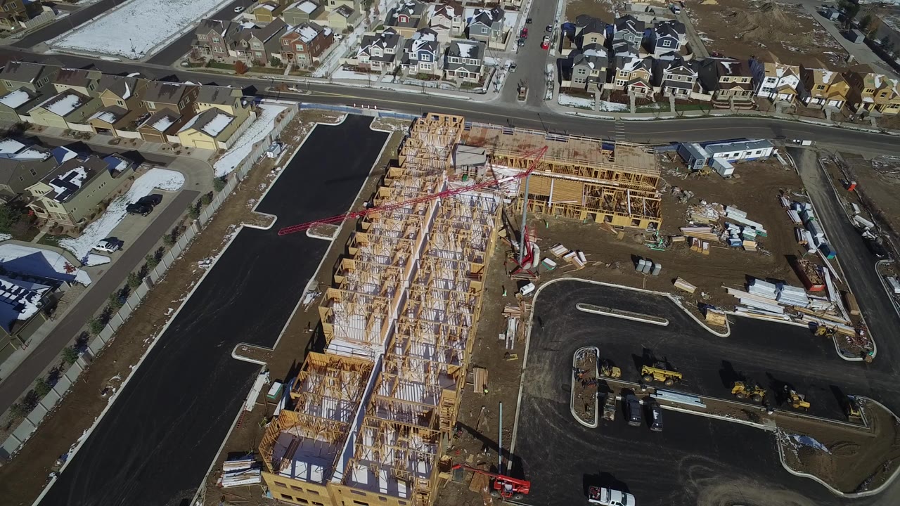 High aerial frame over Hidden Lake, Colorado showing wood framing on a construction site, a dark paved access road, finished homes beyond the street, snow patches, and no visible people.