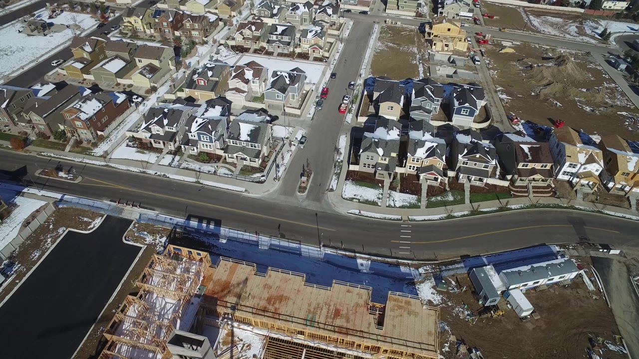Aerial frame in Hidden Lake, Colorado with a broad road crossing the lower frame, rows of private homes above it, a construction slab and framing below it, snow on roofs and yards, and no visible people.
