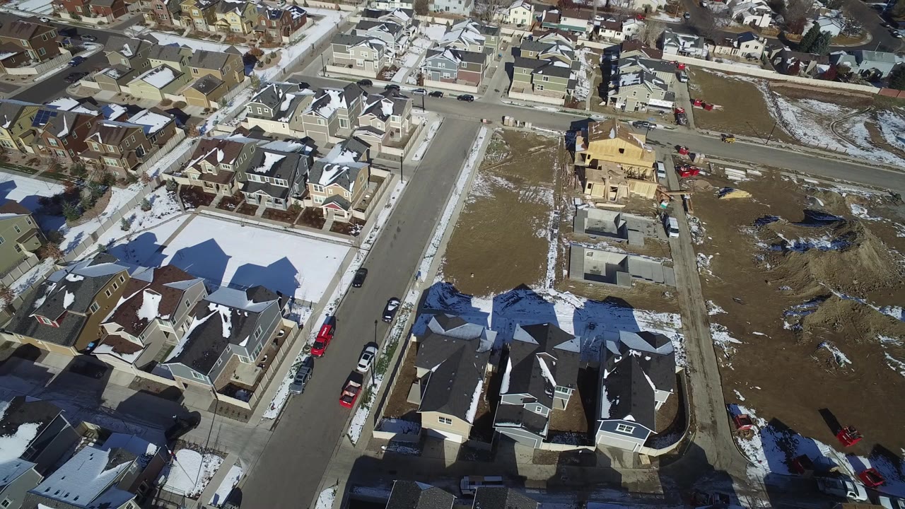 High oblique aerial view over Hidden Lake, Colorado houses, snowy rooftops, a residential road, brown open parcels, and no visible people.