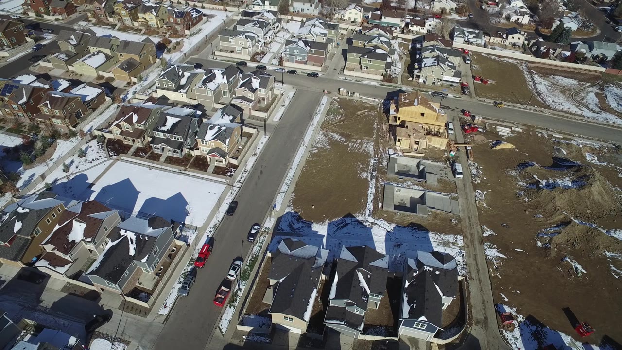 Aerial view in Hidden Lake, Colorado with a partially framed yellow house near the center, surrounding finished homes, open dirt lots, and no visible people.