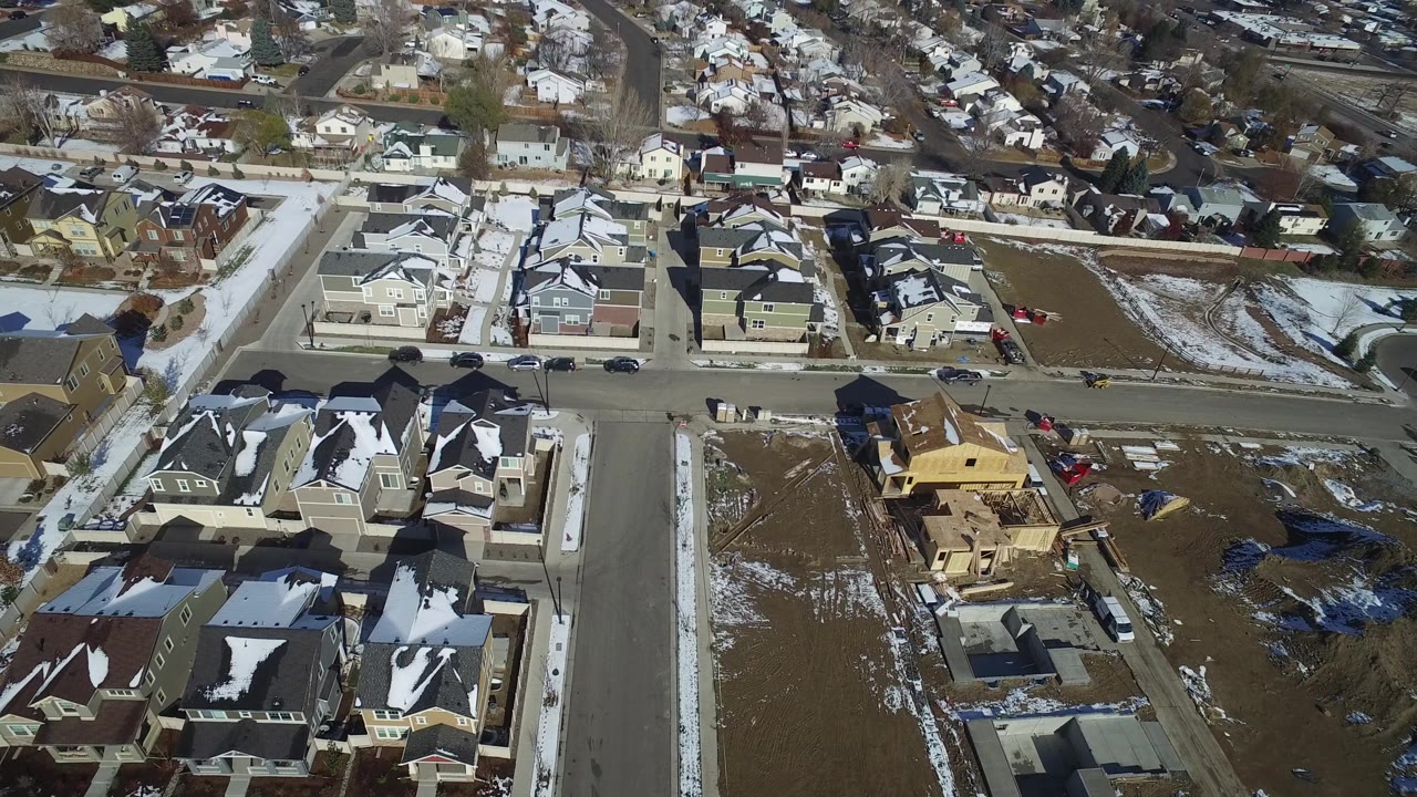 Drone view over Hidden Lake, Colorado with a residential street crossing the frame, clustered houses with snow on roofs, parked cars along the curb, open lots, and no visible people.