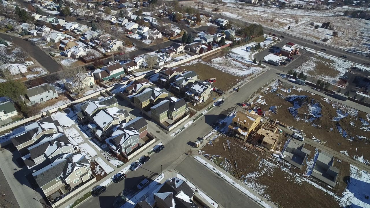 High aerial view over Hidden Lake, Colorado homes and paved neighborhood roads, with open brown lots, patchy snow on roofs and ground, parked cars, and no visible people.