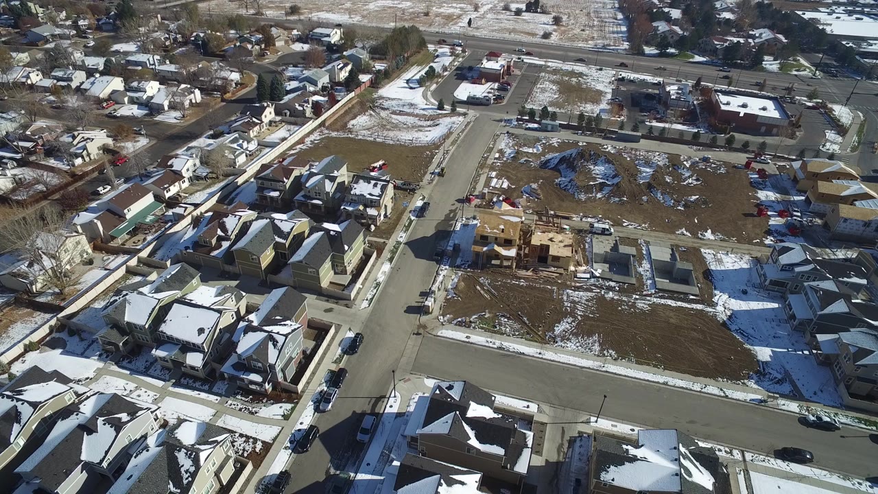 Aerial view of suburban streets, rooftops, open lots, cars, and patchy snow.