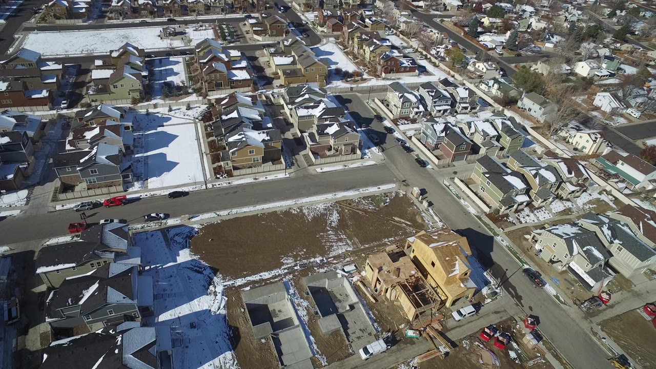Aerial view of suburban homes, streets, open lots, and patchy snow.