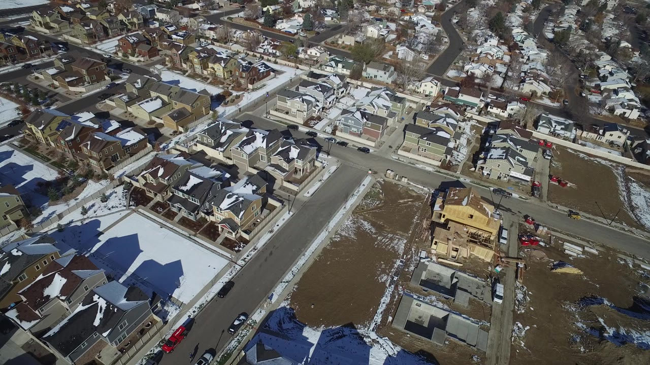 A high aerial frame over Hidden Lake, Colorado shows snow-lined residential streets, finished homes, open dirt lots, and a wood-framed house under construction with no visible people.