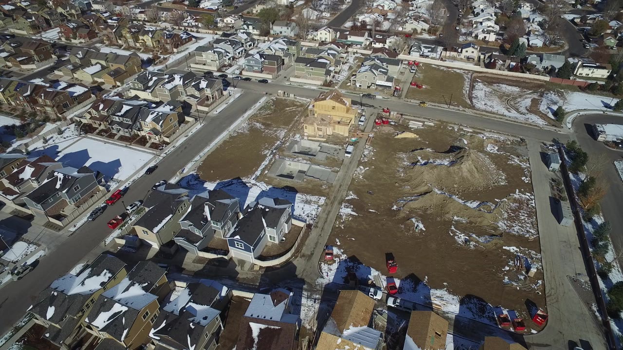 Aerial view of suburban homes, streets, vacant lots, construction piles, and patchy snow.