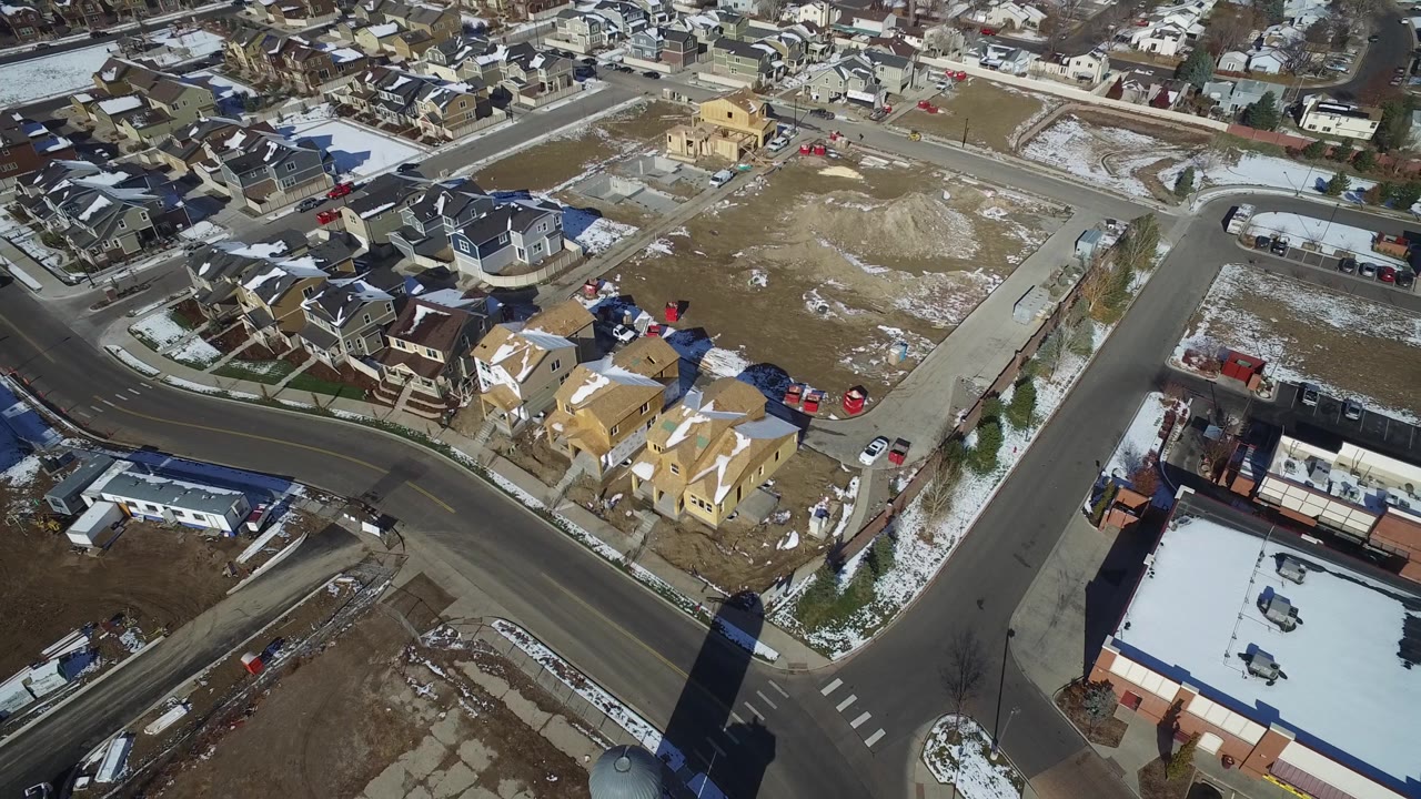 The second Hidden Lake, Colorado frame keeps the curved road central, with rows of houses beyond it, a flat-roof commercial building at lower right, scattered cars, and snow patches, with no visible people.