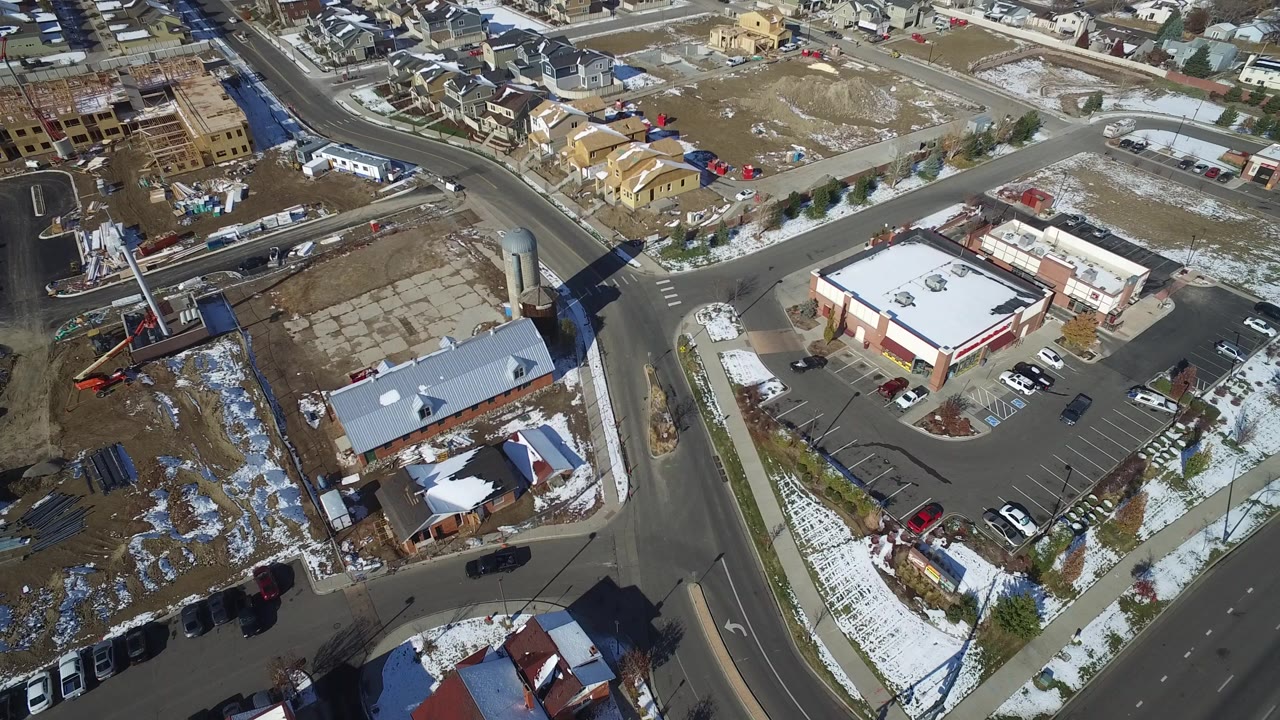 Aerial view of a suburban intersection with roads, parking lots, buildings, cars, and snow patches.