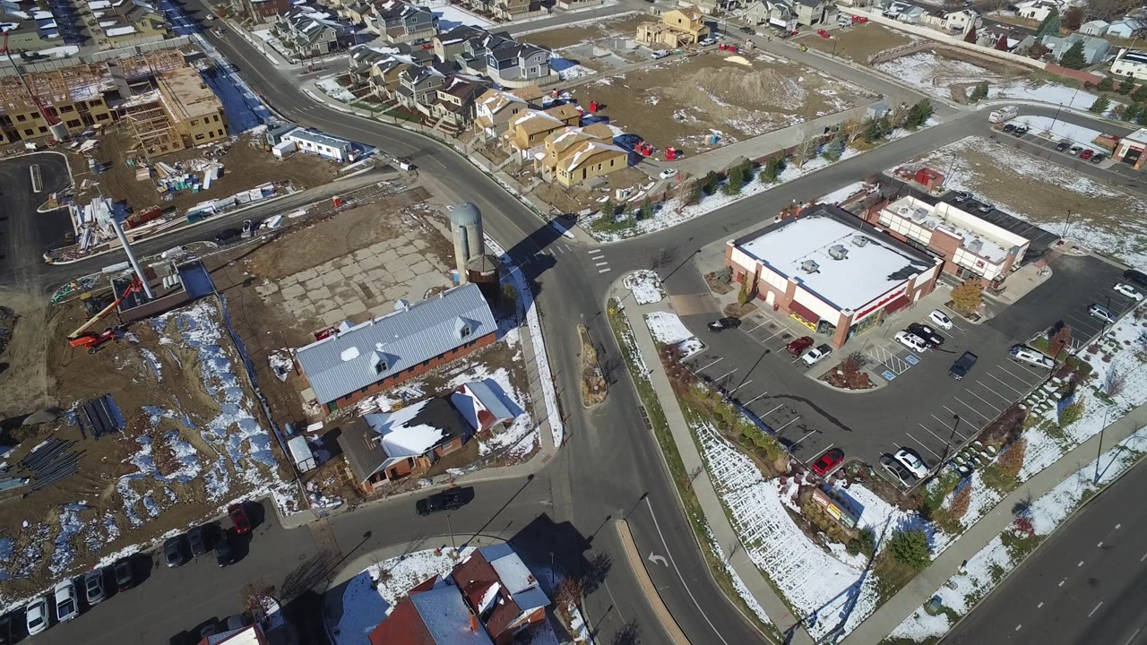 Aerial view in Hidden Lake, Colorado with the road curve and parking lots more centered, showing low retail-style buildings, cars, construction parcels, snow patches, and no visible people.