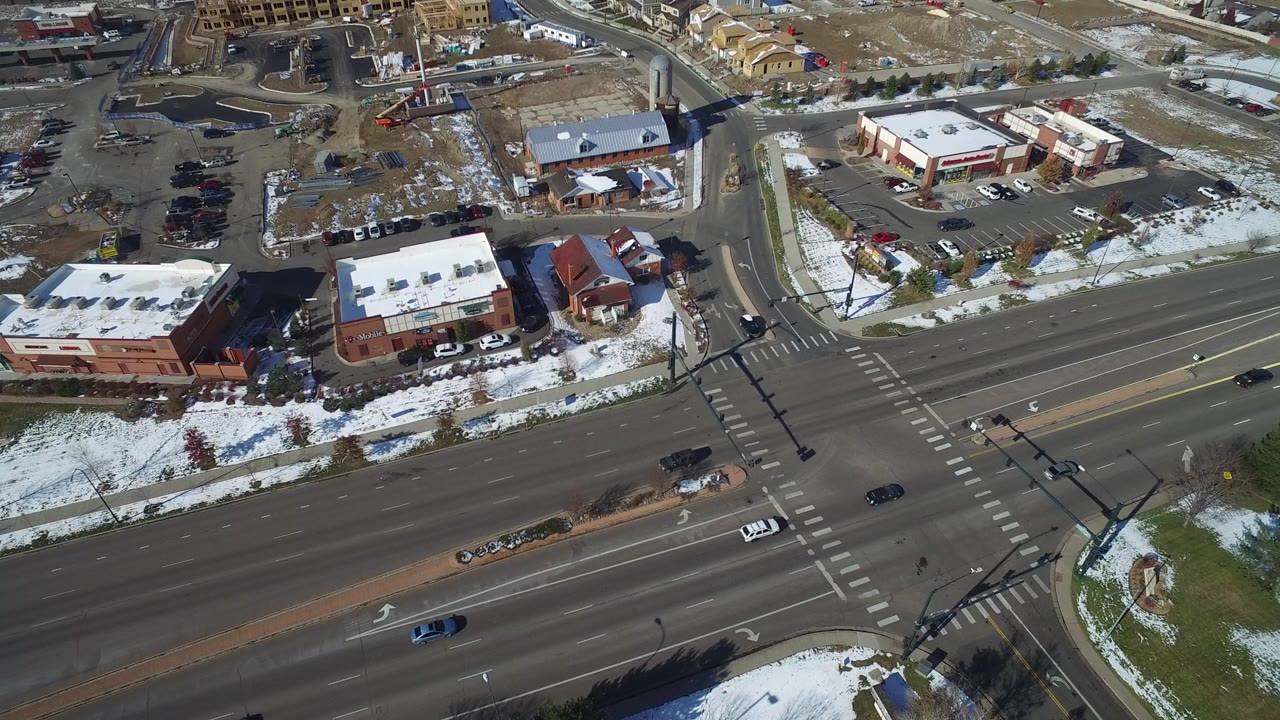 Aerial frame over Hidden Lake, Colorado showing a multi-lane road, crosswalks, low commercial buildings, parking lots, snow patches, and no visible people.