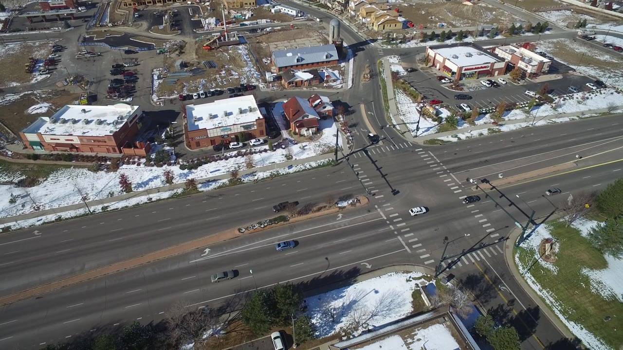Drone view of a wide street intersection with several cars, parking lots, and snowy landscaped areas.