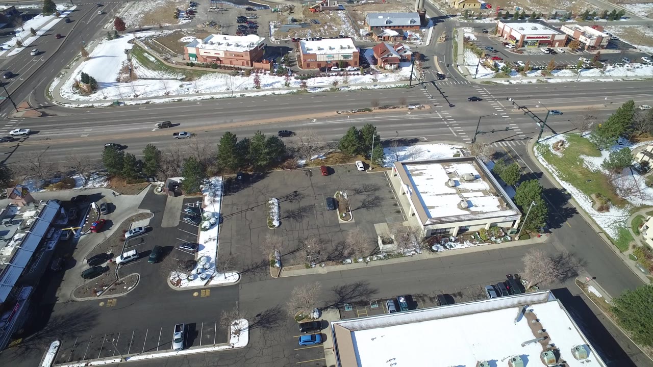 High aerial view in Hidden Lake, Colorado with a broad asphalt lot, a few parked cars, flat commercial roofs, moving road traffic, snow along curbs, and no visible people.