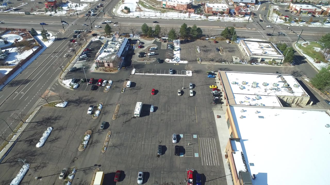 High aerial view in Hidden Lake, Colorado showing a mostly open parking lot, flat-roof commercial buildings, road lanes, snow patches, parked vehicles, and no visible people at the start of the clip.