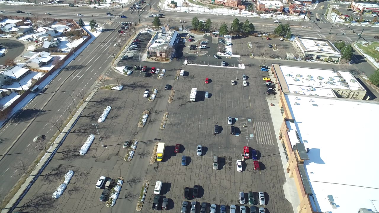 Aerial view of parking rows, a road, buildings, and residential blocks beyond.