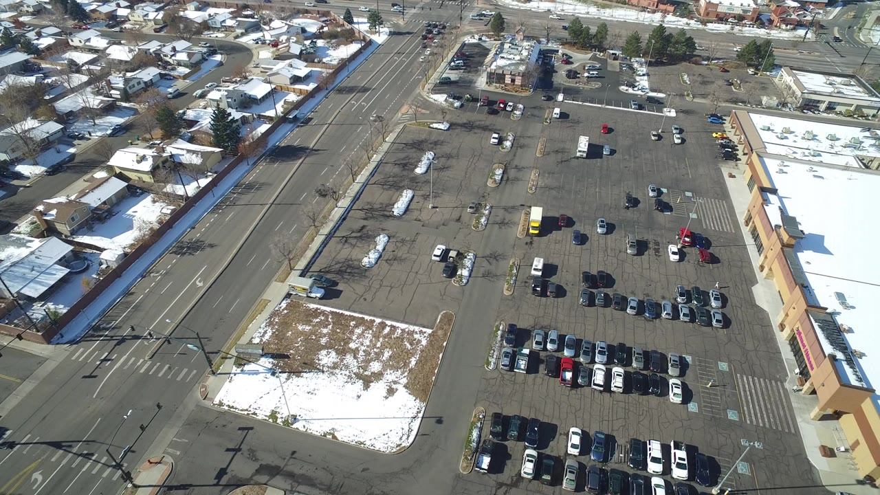 High aerial frame over Hidden Lake, Colorado showing a parking lot with rows of cars, a tan building at right, snowy lawns, and residential streets beyond.