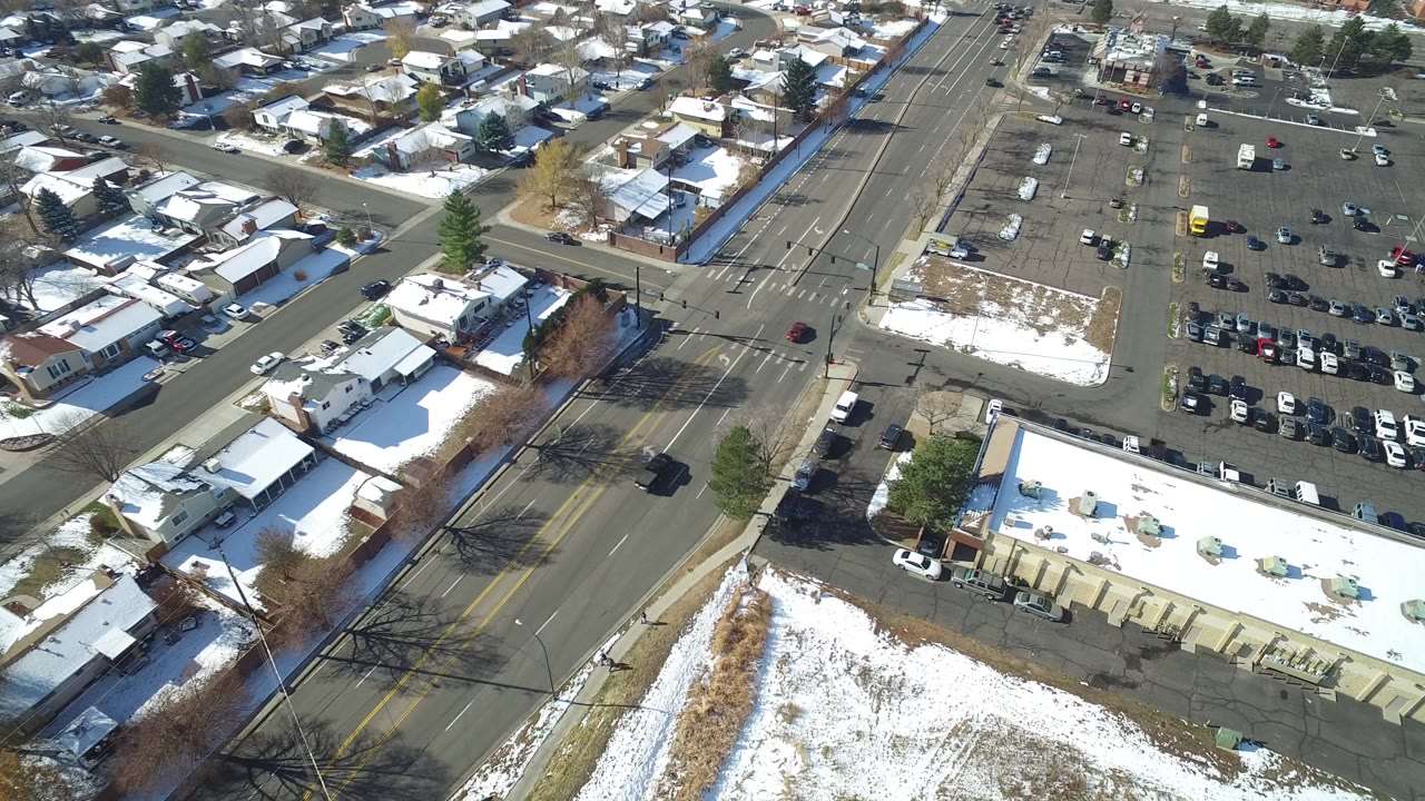 High aerial frame over Hidden Lake, Colorado with snow-covered rooftops, a wide road intersection, parked cars, bare trees, and no visible people.