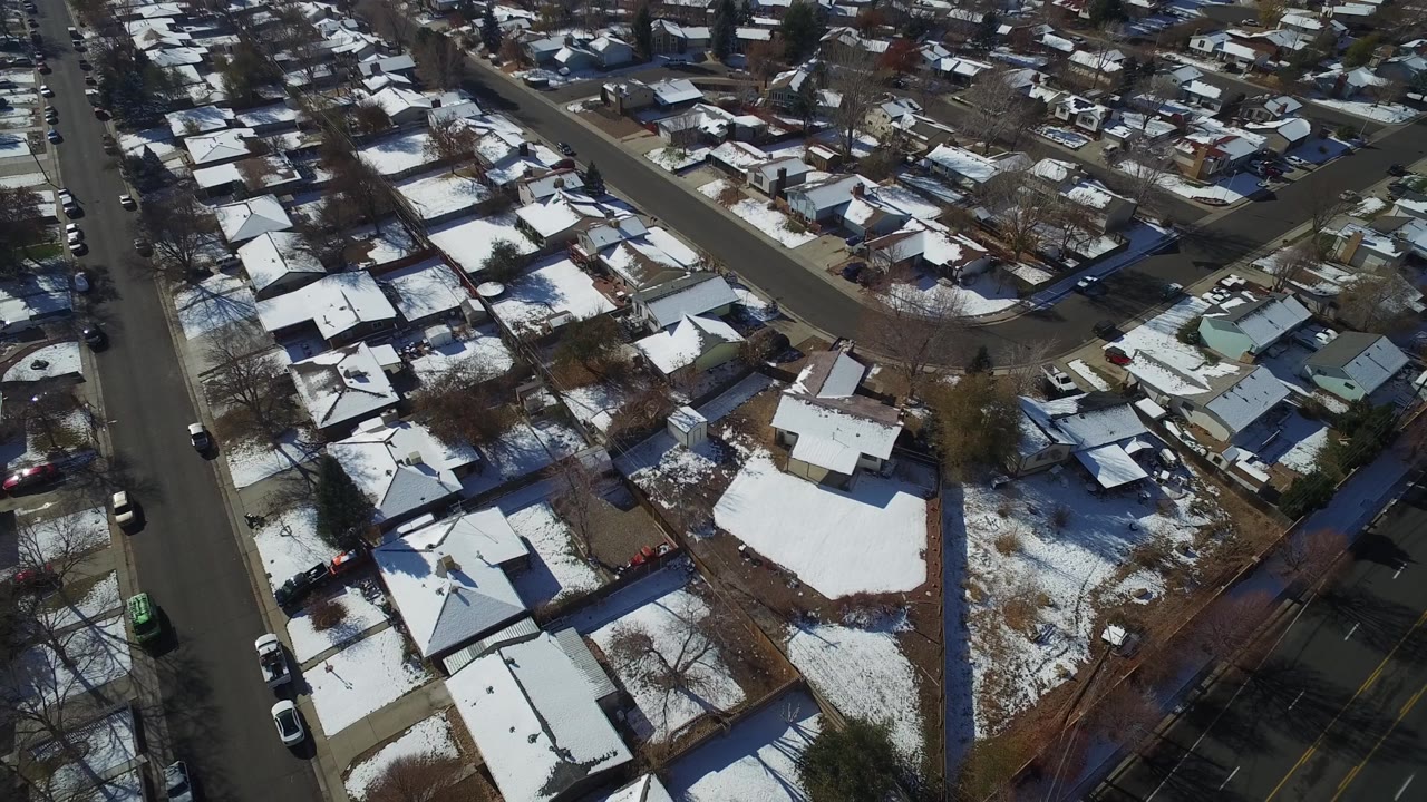 Aerial frame over Hidden Lake, Colorado showing a main road beside residential blocks, white rooftops, winter yards, parked vehicles, and no visible people.